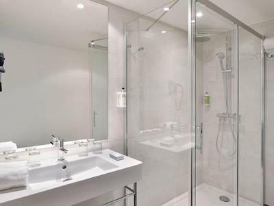 Moderní koupelna se skleněným sprchovým koutem, umyvadlem a vysoušečem vlasů v bílé a šedé.