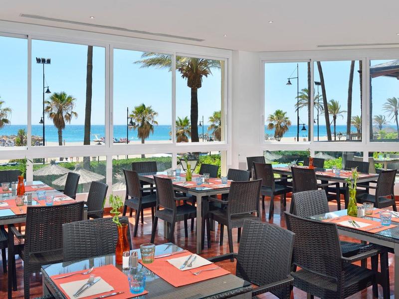 Moderní restaurace s výhledem na moře, velkými okny a tmavými židlemi.