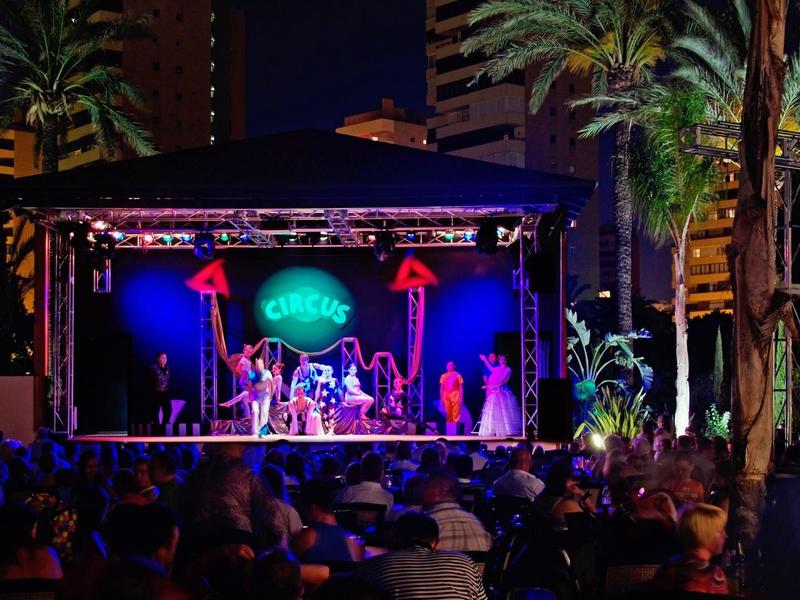 Koncert na jevišti s barevnými světly a davem venku v noci.