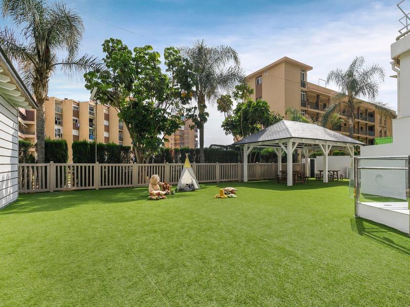 Velká travnatá plocha s altánem a dětským hřištěm vedle hotelových budov pod modrou oblohou.