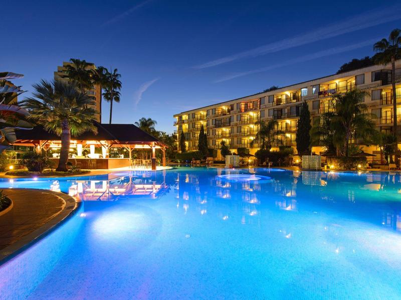 Osvětlený hotelový bazén v noci s přilehlou budovou a palmami.