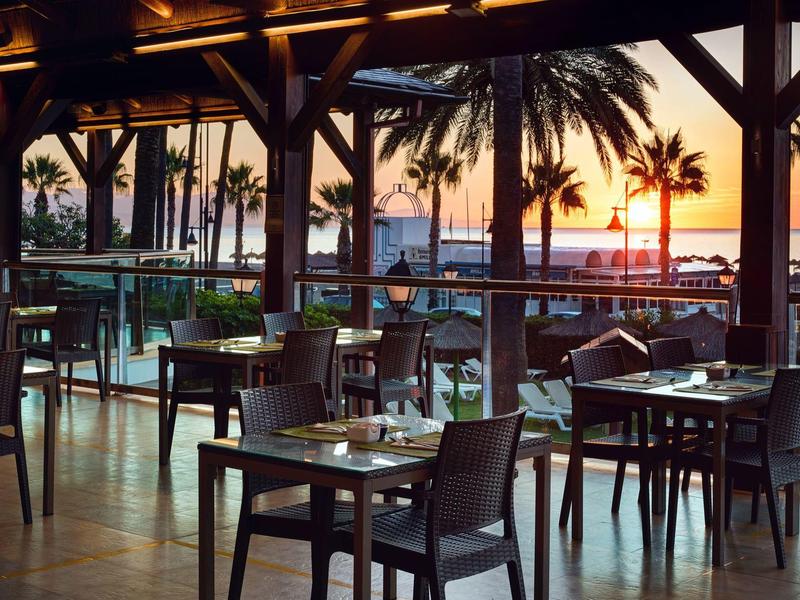 Restaurace s výhledem na palmy a západ slunce nad vodou.