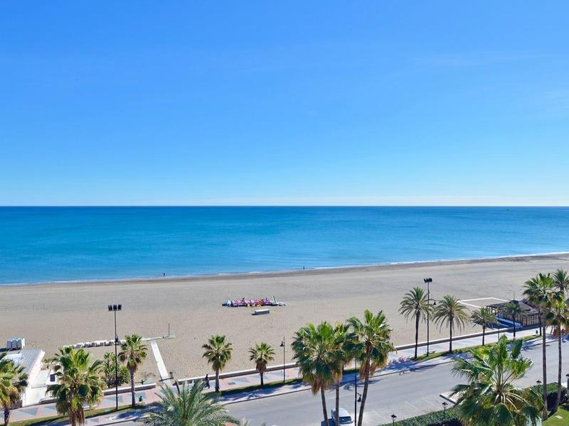 Široká písečná pláž s palmami podél pobřežní silnice pod jasně modrou oblohou.