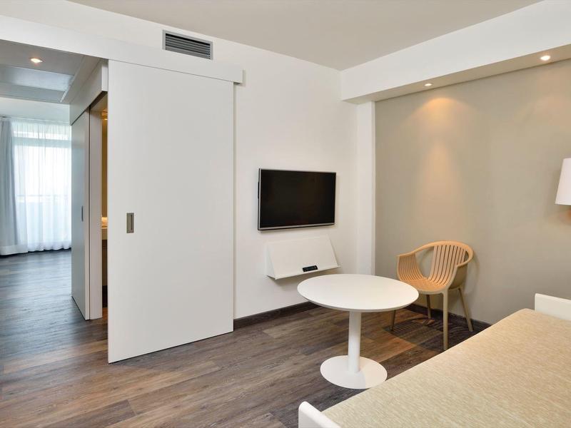 Moderní, světlý hotelový pokoj s plochou TV, stolem, židlí a teplou dřevěnou podlahou.