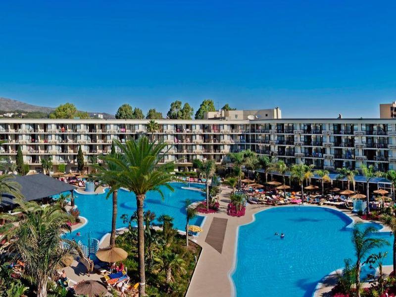 Velký hotel s bazénem a mnoha lehátky pod modrou oblohou.