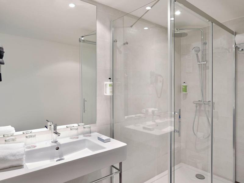 Moderní koupelna se skleněným sprchovým koutem, umyvadlem a vysoušečem vlasů v bílé a šedé.