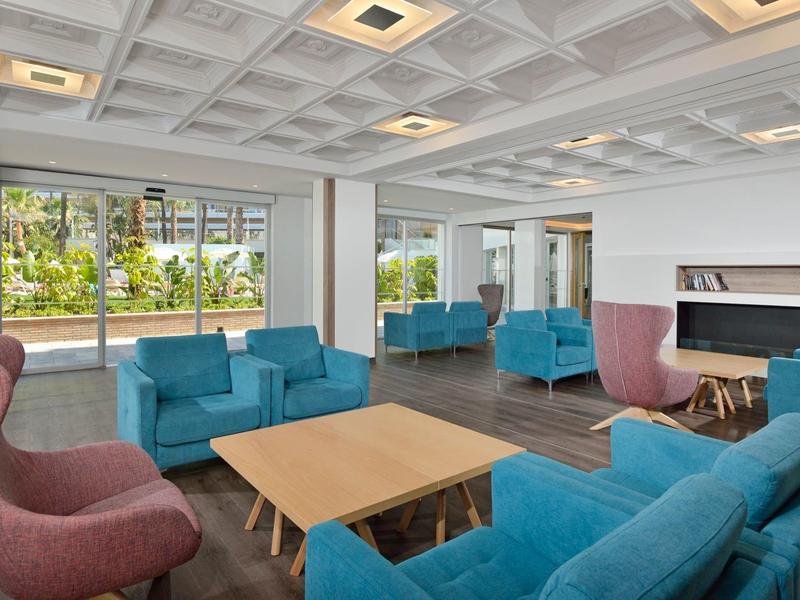 Heller Wohnraum mit blauen Sofas, rosa Sesseln, Holztisch und großen Fenstern mit Aussicht auf Bäume.