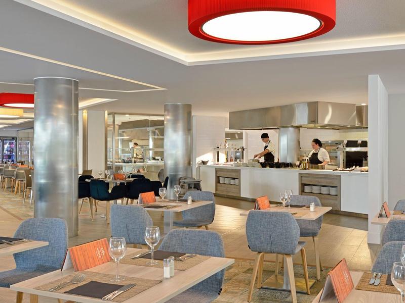 Modernes Restaurant mit hellen Holztischen, grauen Stühlen und roten Lampen an der Decke.