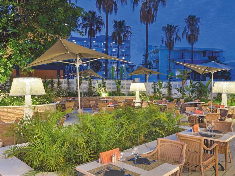 Außenbereich eines Restaurants mit Tischen, Stühlen, Sonnenschirmen und Palmen bei Abenddämmerung.