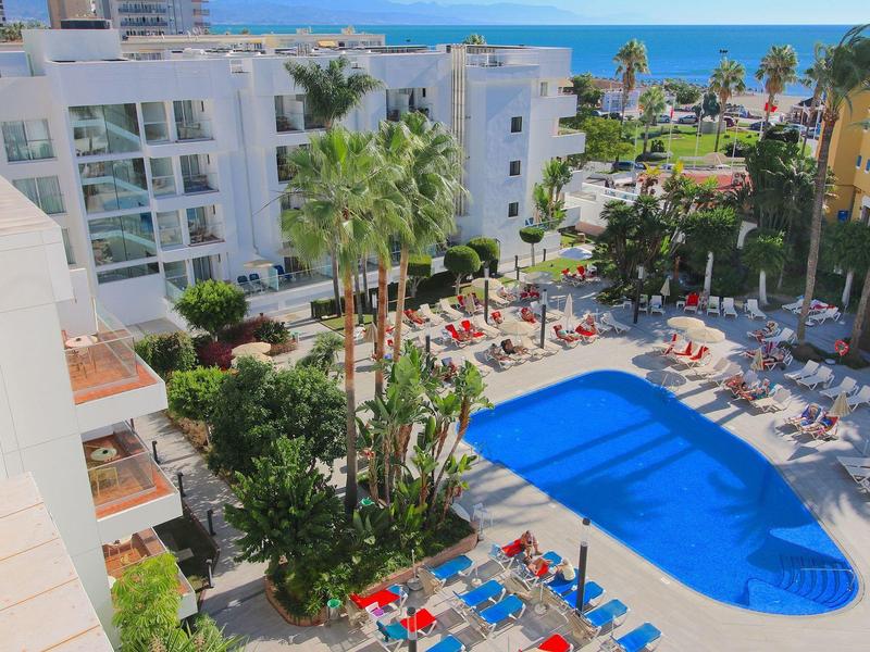 Hotel mit blauem Pool, Liegestühlen und Palmen, Meer im Hintergrund bei sonnigem Himmel.