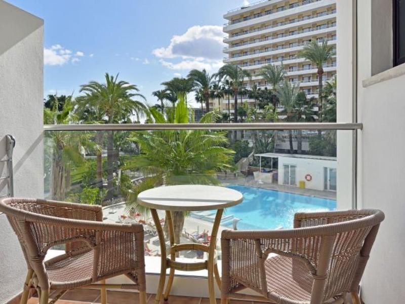 Balkon mit zwei Korbstühlen und Tisch, Blick auf blauen Pool und grüne Palmen bei blauem Himmel.