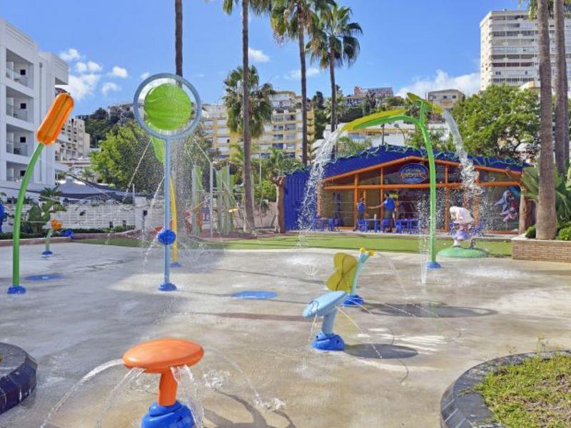 Bunter Wasserspielplatz mit Spritzduschen, Palmen und Hotelgebäuden im Hintergrund.