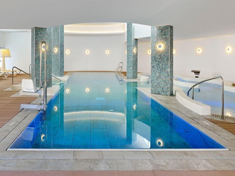 Moderner Innenpool mit klar blauem Wasser, Holzterrasse, Liegestühlen und warmem Licht an den Wänden.