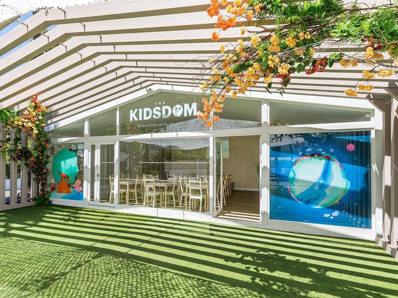 Moderner, heller Eingangsbereich mit Glasfront, Beschattung und Kunstrasen, umgeben von Pflanzen.