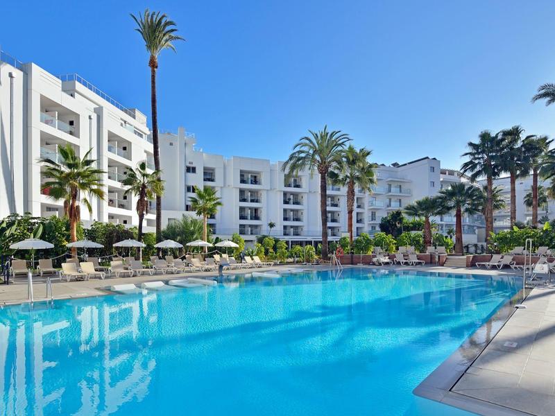 Großer blauer Pool vor einem weißen Hotelgebäude, umgeben von Palmen und Sonnenschirmen.