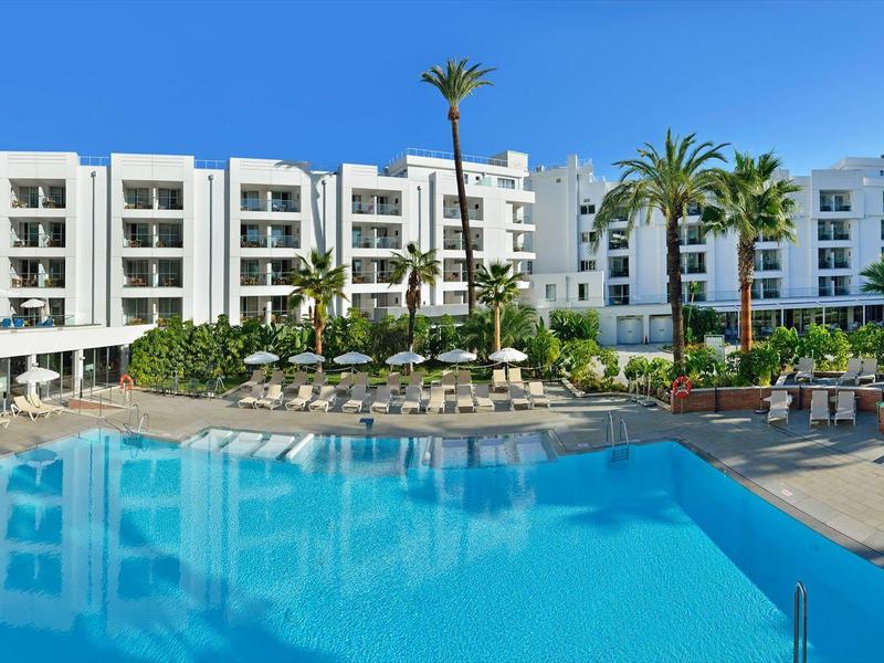 Großer blauer Pool vor weißem mehrstöckigem Hotel mit Palmen und Sonnenschirmen.