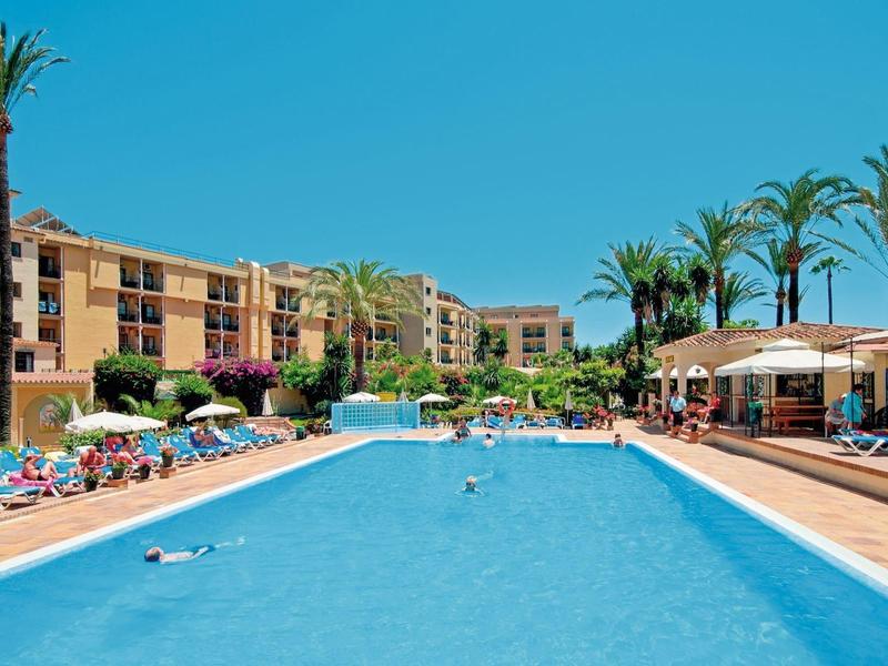 Großer blauer Pool mit Liegestühlen, Palmen und mehrstöckigem Hotel bei klarem Himmel.