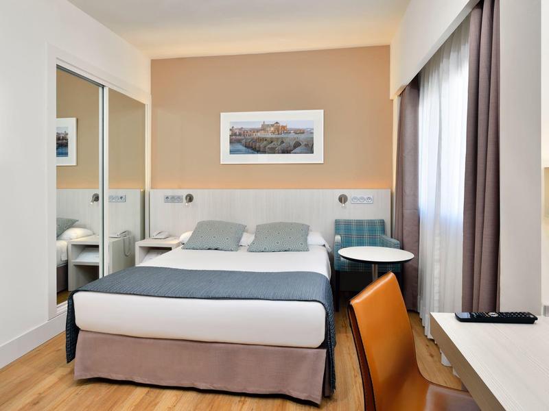 Modernes Hotelzimmer mit Doppelbett, Schreibtisch, Stuhl, Spiegel und Fenster mit Vorhängen.