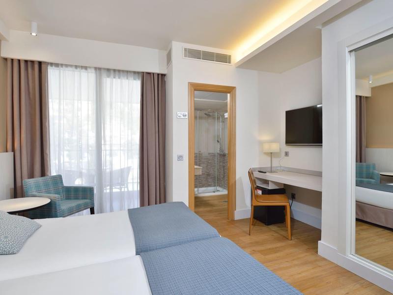 Modernes Hotelzimmer mit zwei Betten, großem Spiegel, Fernseher und Balkon mit Vorhängen.