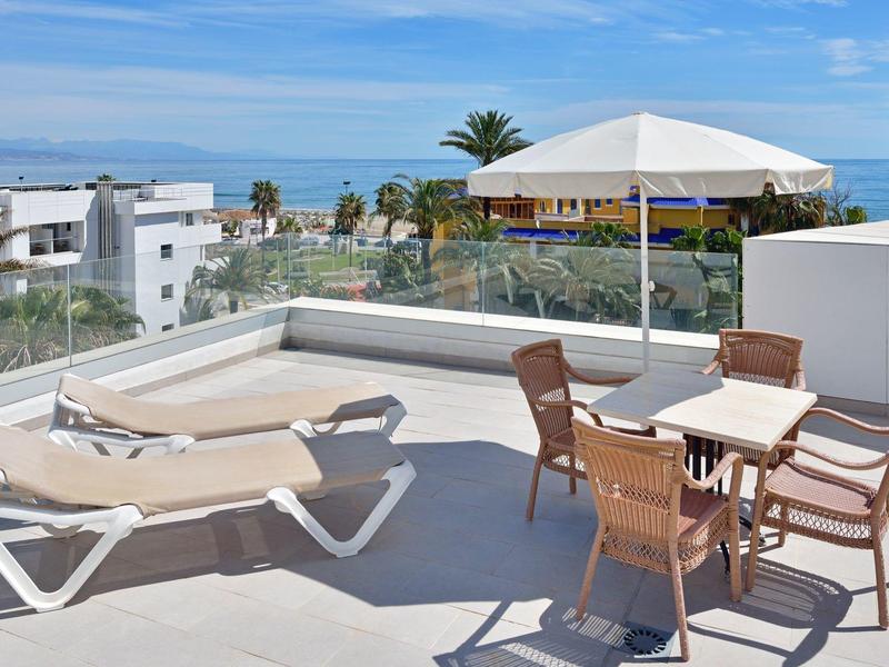 Dachterrasse mit Sonnenschirm, Tisch, Stühlen und Liegestühlen, Blick auf Meer und Palmen.