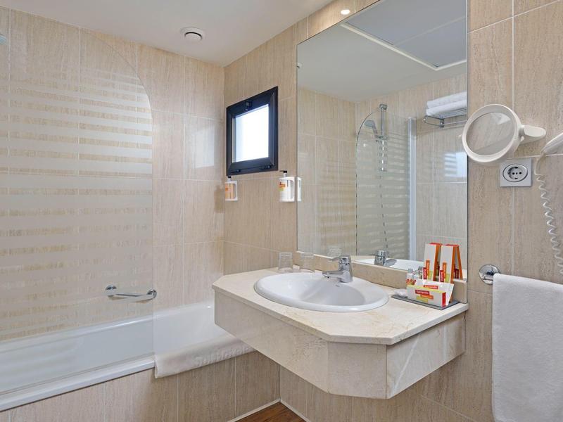 Hell beleuchtetes Badezimmer mit Badewanne, großem Spiegel, Waschbecken und Handtuchhalter.