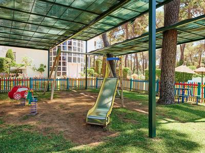 Venkovní hřiště s klouzačkou a houpačkou pod zeleným přístřeškem obklopené plotem.