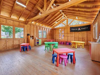 Jasná dřevěná místnost s barevnými dětskými stolky a stoličkami, velká okna a stropní ventilátor.