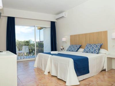 Světlý hotelový pokoj s manželskou postelí, modrými doplňky a balkonem s výhledem ven.