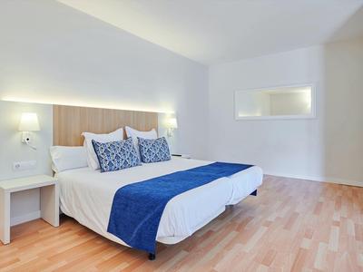 Moderní hotelový pokoj s manželskou postelí, modrým ložním prádlem, dřevěnou podlahou a bílými stěnami.