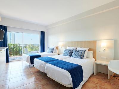 Helles Hotelzimmer mit zwei Betten, blauen Akzenten und Balkon mit Meerblick.