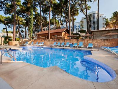 Ein klarer blauer Hotelpool mit Sonnenliegen unter schattigen Bäumen und Gebäuden im Hintergrund.