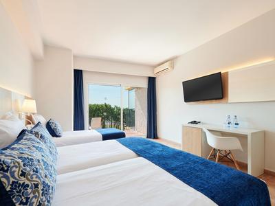 Helles Hotelzimmer mit zwei Betten, Schreibtisch, Fernseher und Blick auf grüne Bäume.