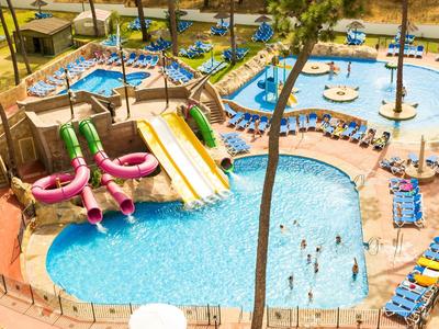 Areál bazénu resortu s vodními skluzavkami, palmami a lehátky za slunečného počasí.