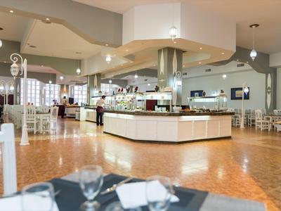 Prostorná hotelová restaurace s bílým nábytkem a centrálním bufetem pod teplým osvětlením.