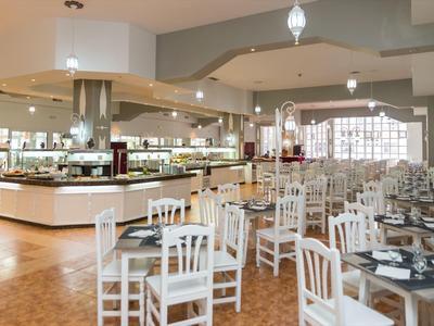 Prostorná hotelová jídelna s bílými židlemi, prostřenými stoly a bufetovými pulty při tlumeném osvětlení.