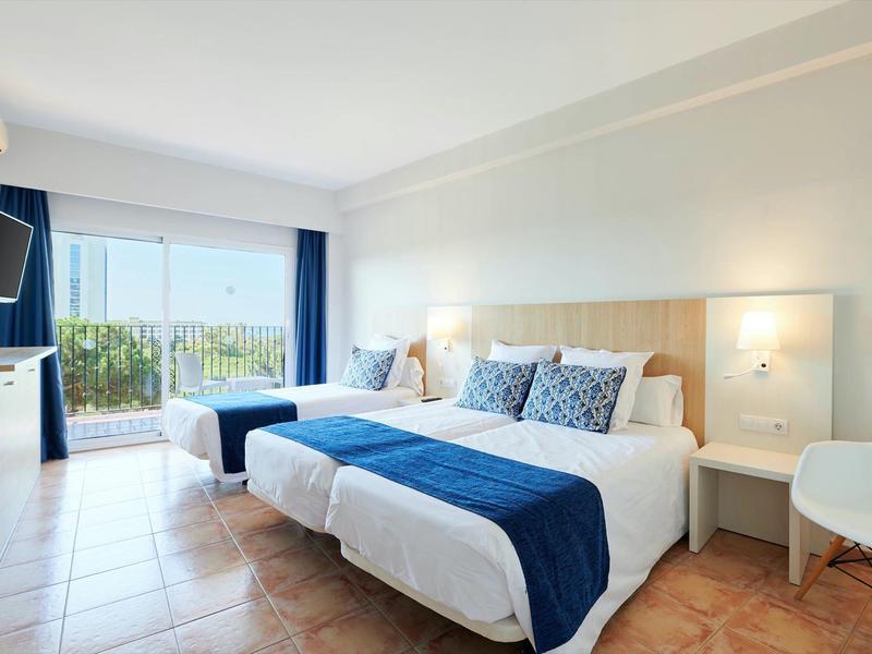 Světý hotelový pokoj s dvěma postelemi, modrými doplňky, terasou a výhledem na zahradu.