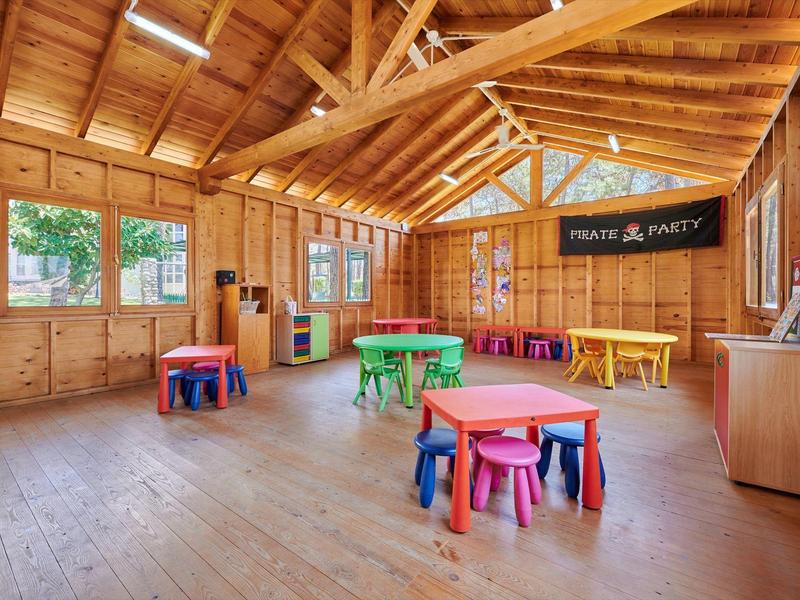 Jasná dřevěná místnost s barevnými dětskými stolky a stoličkami, velká okna a stropní ventilátor.