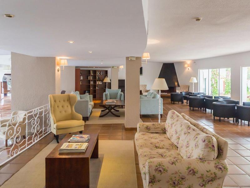 Prostorný hotelový salonek s béžovými pohovkami, křesly a velkými okny vpouštějícími přirozené světlo.