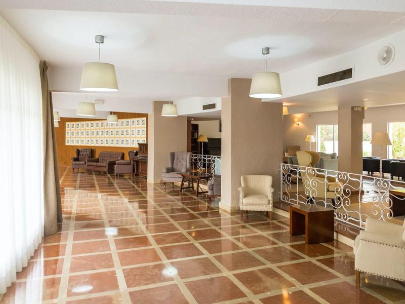 Prostorná hotelová hala s dlažbou, béžovým nábytkem a velkými okny se záclonami.