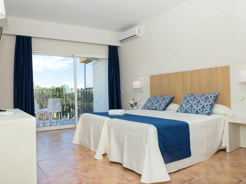 Světlý hotelový pokoj s manželskou postelí, modrými doplňky a balkonem s výhledem ven.