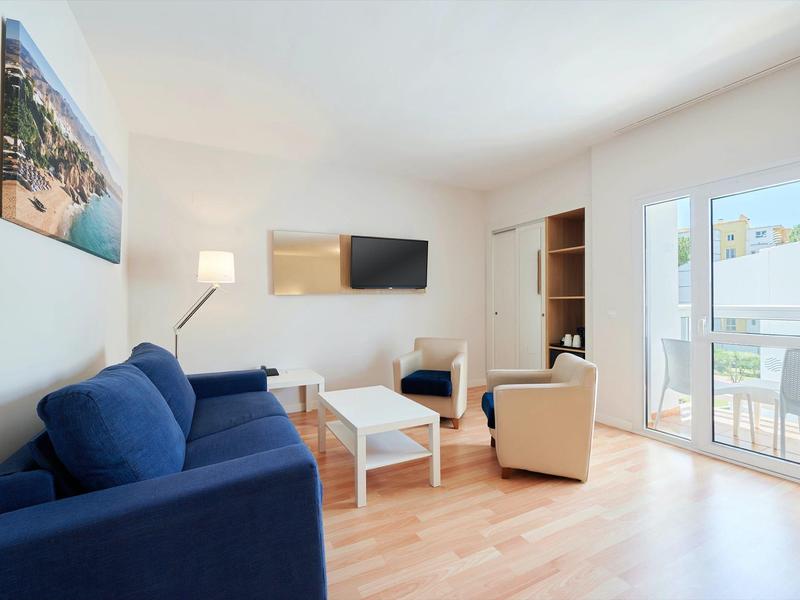 Světý hotelový pokoj s dřevěnou podlahou, modrou pohovkou, béžovým křeslem a přístupem na balkon.