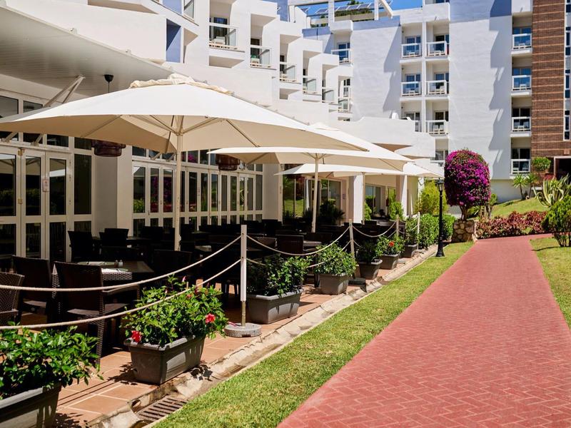 Venkovní posezení s stolky pod velkými slunečníky vedle červené cesty a bílé budovy.