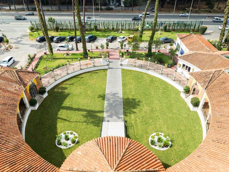 Pohled na kruhovou zahradu se zelenou trávou a cestičkami, obklopenou budovami s terakotovými střechami.