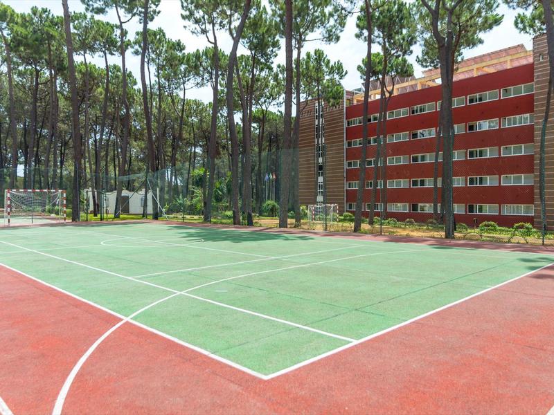 Venkovní basketbalové hřiště vedle červené hotelové budovy obklopené vysokými borovicemi.