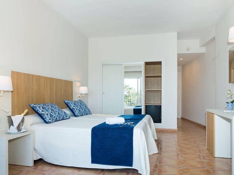 Moderní hotelový pokoj s manželskou postelí, modrými doplňky, nástěnnými lampami, TV a malou ledničkou.