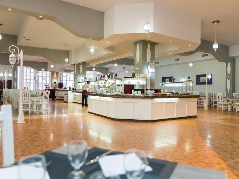 Prostorná hotelová restaurace s bílým nábytkem a centrálním bufetem pod teplým osvětlením.