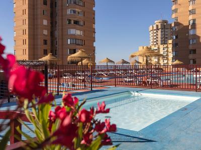 Poolbereich mit Sonnenschirmen und Gebäuden im Hintergrund an einem sonnigen Tag.