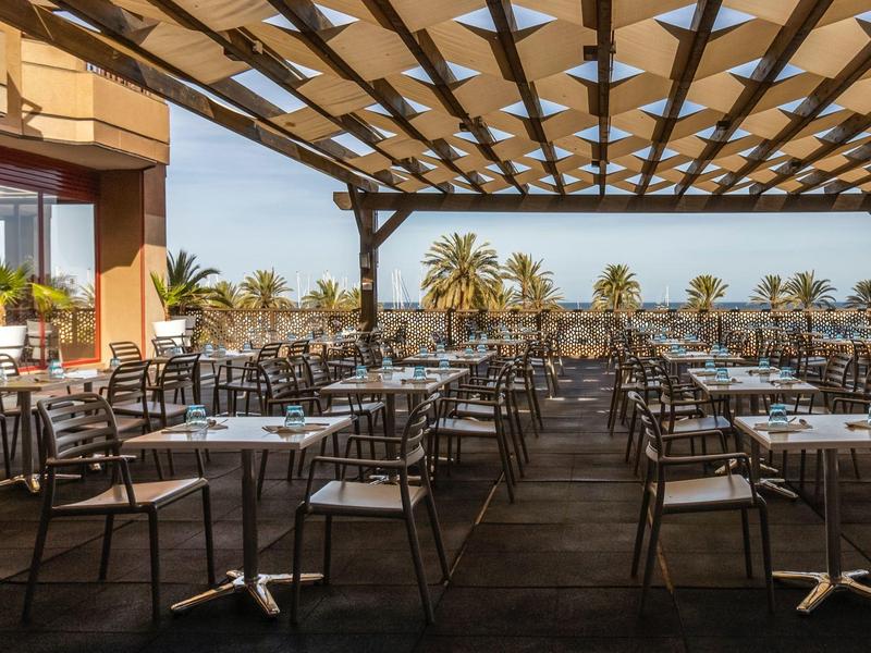 Terrazza coperta con molti tavoli e sedie, vista su palme e mare.