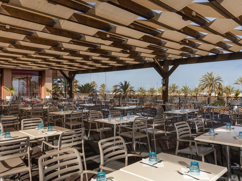 Terrazza coperta di un ristorante con tavoli e sedie, palme sullo sfondo.