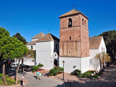 Church with brown tower and white walls under clear blue sky, surrounded by trees.
